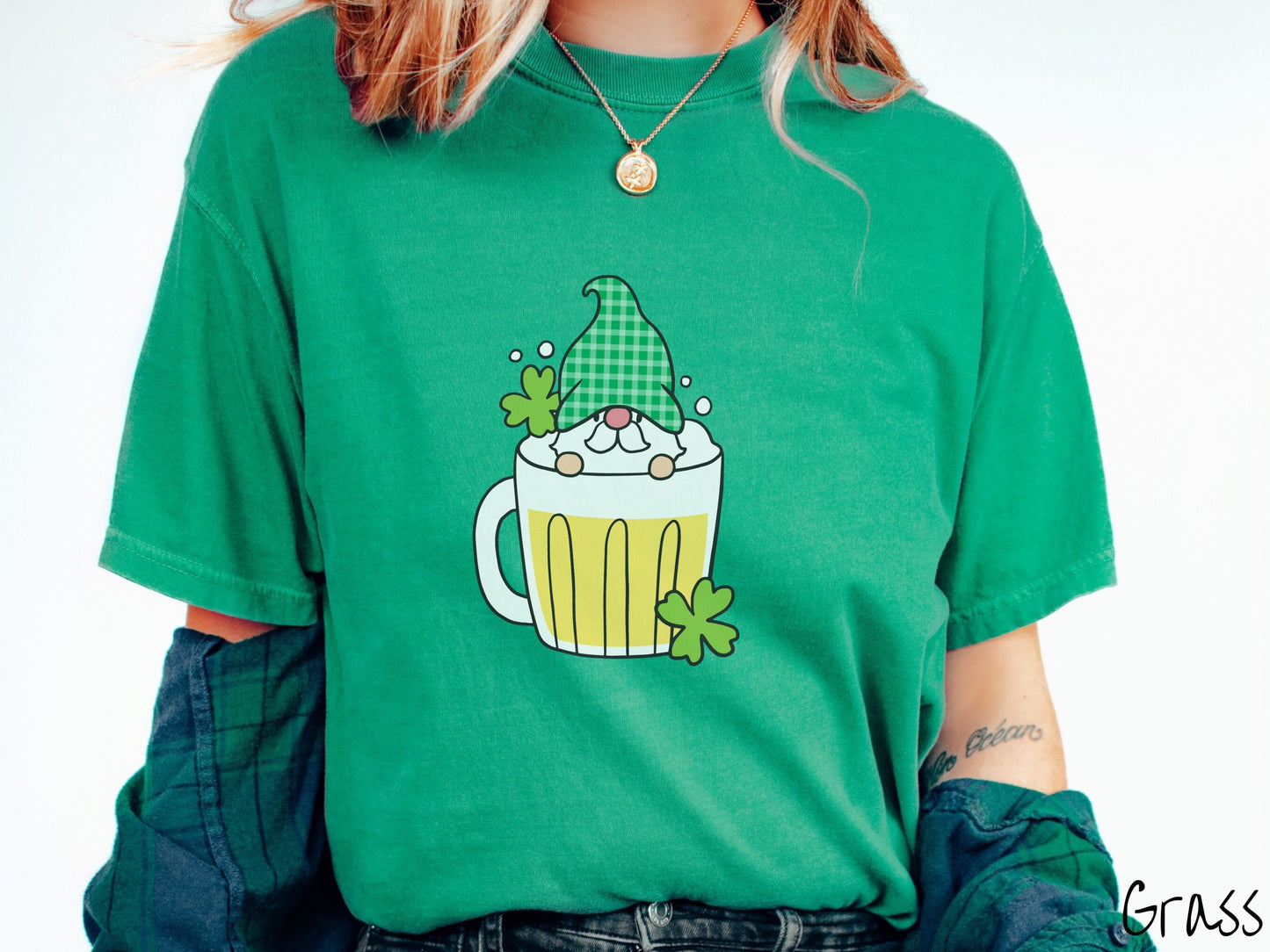 A woman wearing a vintage, grass colored shirt with a beer filled mug in the center with a green hat wearing gnome peeking over the top of the glass and green clovers in the background.