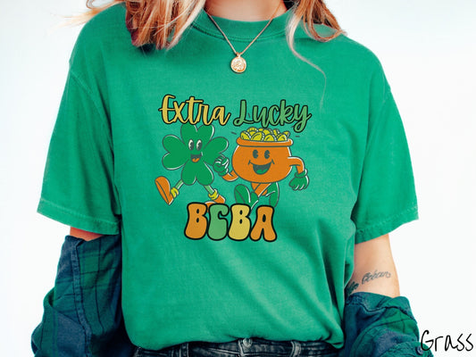 A woman wearing a vintage, grass colored shirt with the text Extra Lucky BCBA in yellow, orange, and green font. Between the text are a green clover and orange pot of gold smiling and walking together in step.