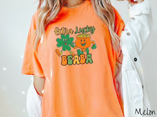 A woman wearing a vintage, melon colored shirt with the text Extra Lucky BCABA in yellow, orange, and green font. Between the text are a green clover and orange pot of gold smiling and walking together in step.