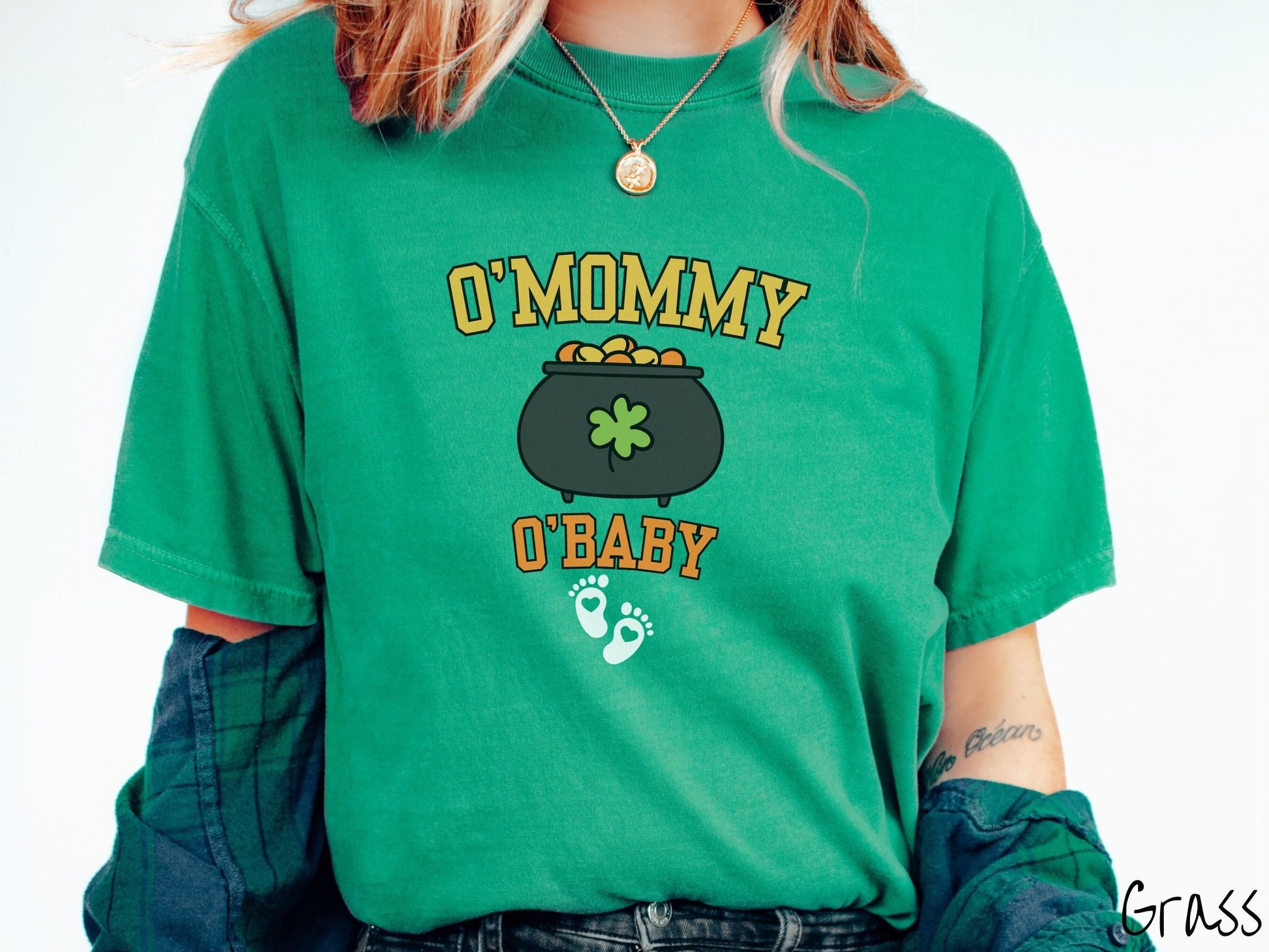 A woman wearing a vintage, grass colored shirt with text O Mommy O Baby in yellow and orange font, a black pot of gold with a green clover on it and yellow and orange gold coins inside. Below all this are two white baby footprints with pink hearts.