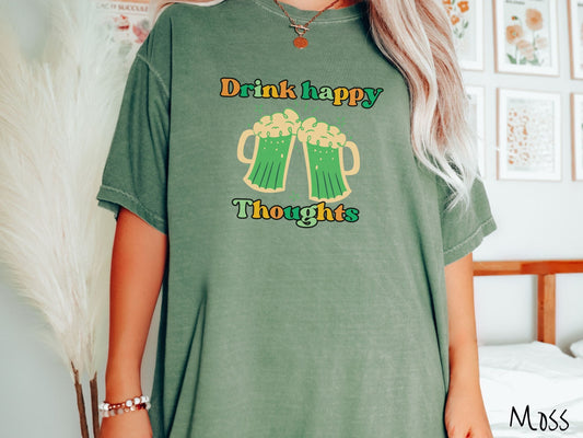 A woman wearing a vintage, moss colored shirt with orange, green, and yellow text Drink Happy Thoughts, between the text are two green beer glasses with beer foaming over the tops clinking together cheering.