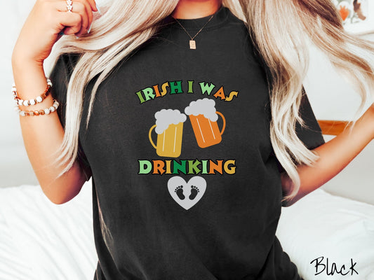 A woman wearing a vintage, black colored shirt with text Irish I Was Drinking in green, orange and yellow fonts. In between the text are two yellow and orange mugs of foaming beer clinking together, and below it is a heart with two baby feet inside.