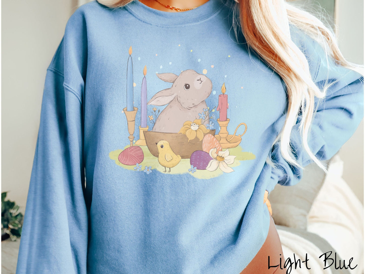 A woman wearing a cute, vintage light blue colored sweatshirt with a gray bunny rabbit sitting in between three lit candles, some red, orange, and purple eggs, and yellow and white flowers.
