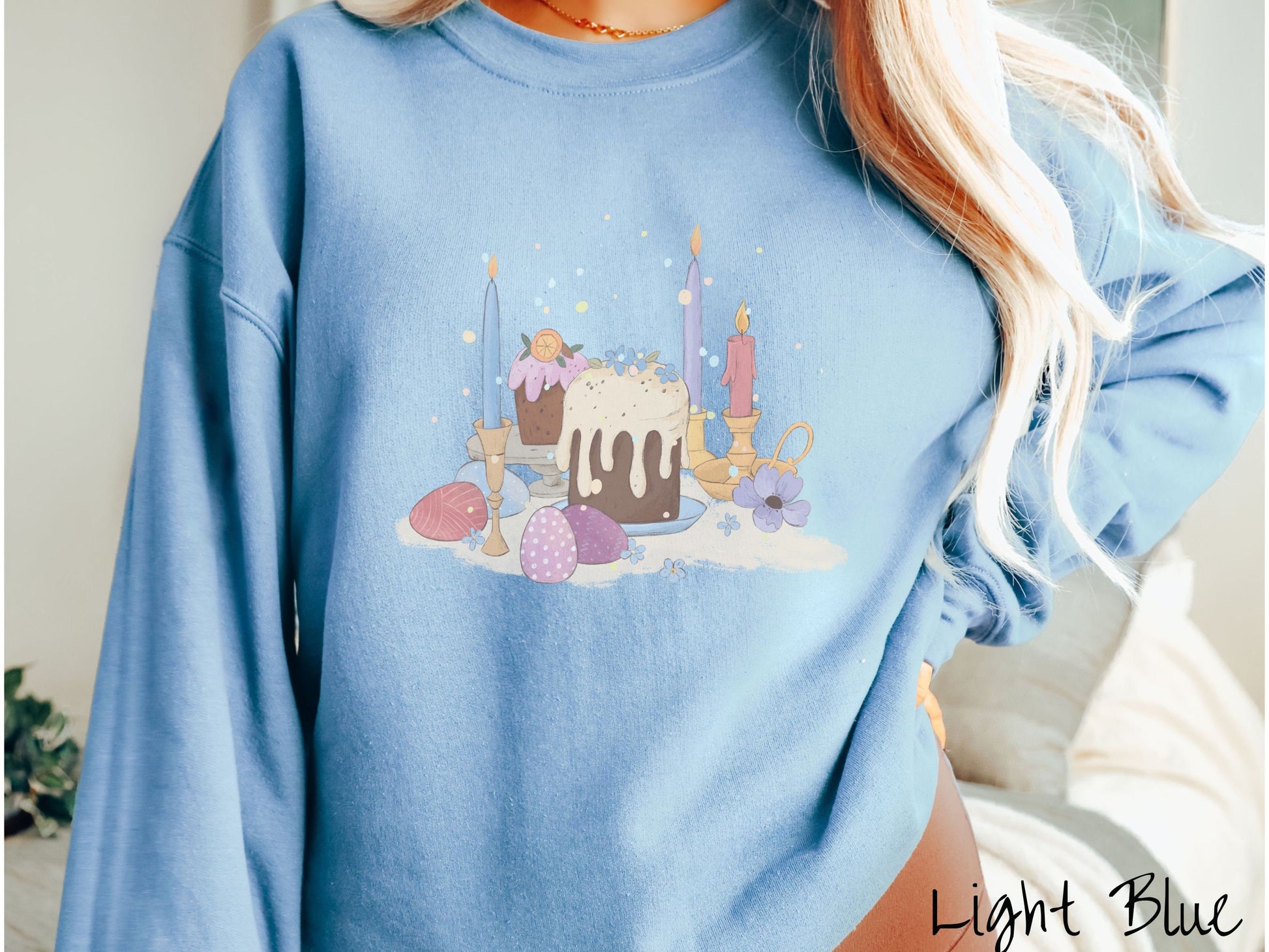 A woman wearing a cute, vintage light blue colored sweatshirt with an assortment of colorful candles with wax running down the sides in the center, scattered around the candles are pink, purple, red, and blue colored Easter eggs.