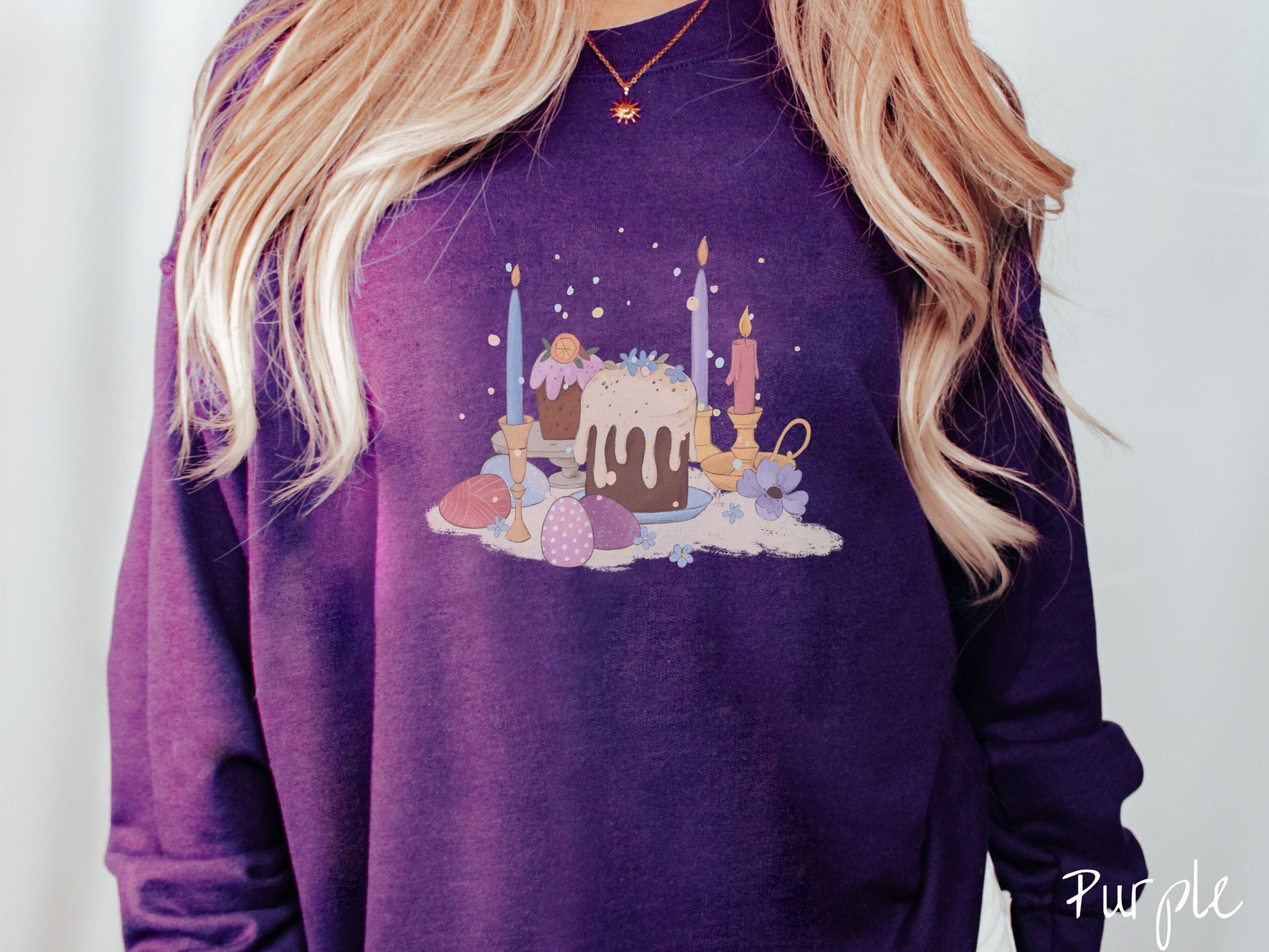 A woman wearing a cute, vintage purple colored sweatshirt with an assortment of colorful candles with wax running down the sides in the center, scattered around the candles are pink, purple, red, and blue colored Easter eggs.