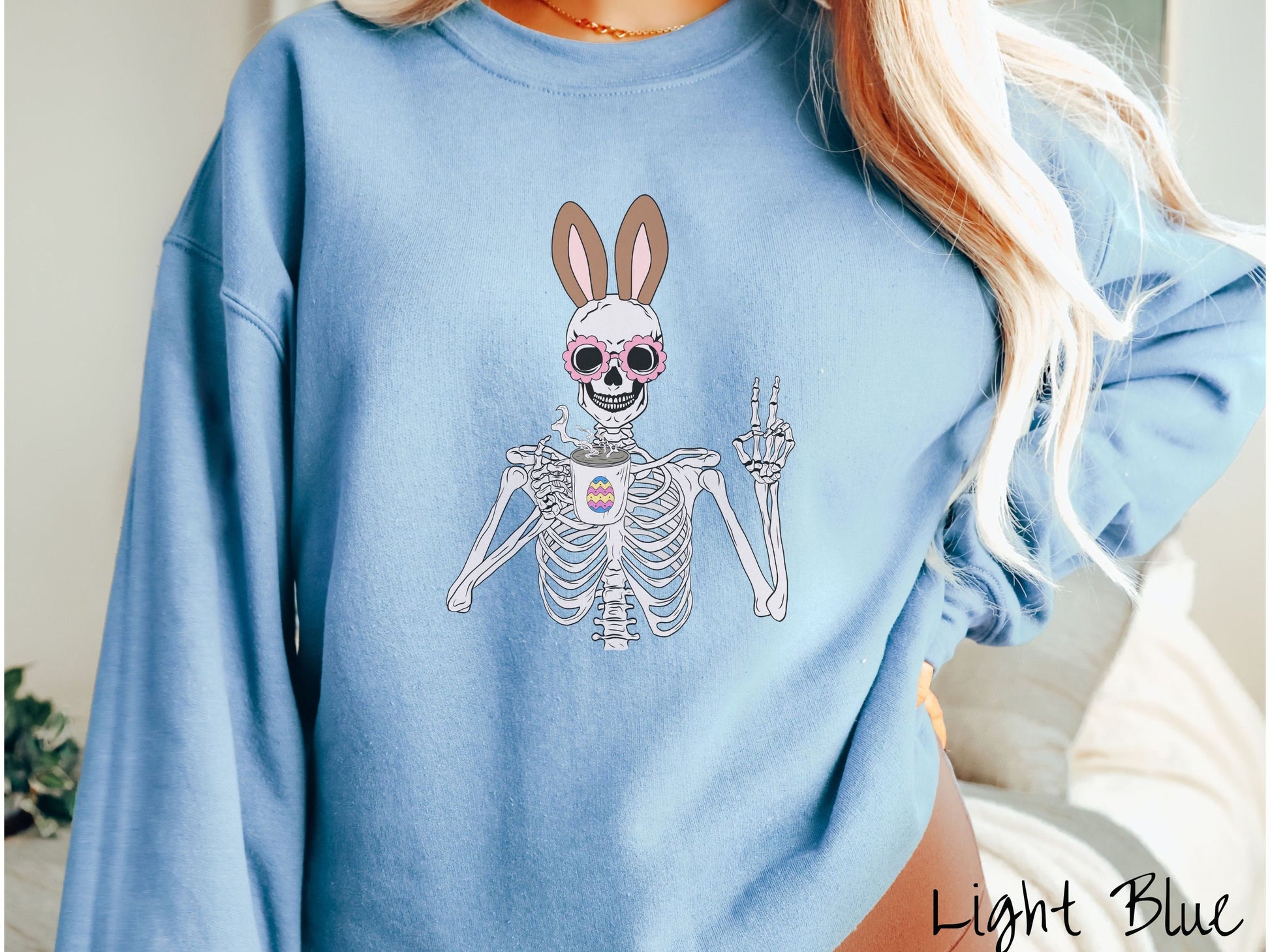 A woman wearing a cute, vintage light blue colored sweatshirt with a pink-toned skeleton wearing pink glasses and brown bunny ears, holding a cup of coffee and holding up a peace sign with the other hand. The coffee cup has a colorful Easter egg.