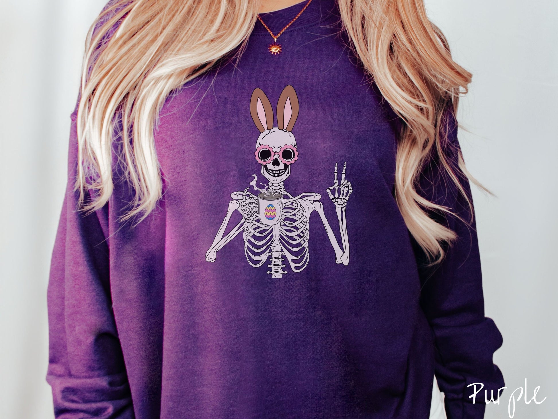 A woman wearing a cute, vintage purple colored sweatshirt with a pink-toned skeleton wearing pink glasses and brown bunny ears, holding a cup of coffee and holding up a peace sign with the other hand. The white coffee cup has a colorful Easter egg.