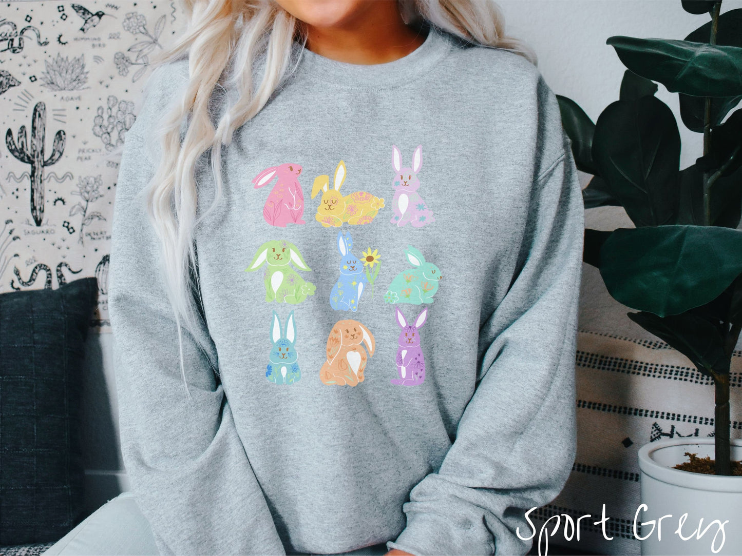 A woman wearing a cute, vintage sport grey colored sweatshirt with a three by three grid of colorful bunny rabbits. They are from left to right pink, yellow, purple, green, blue, seafoam, light blue, orange, and purple with flowers near them.