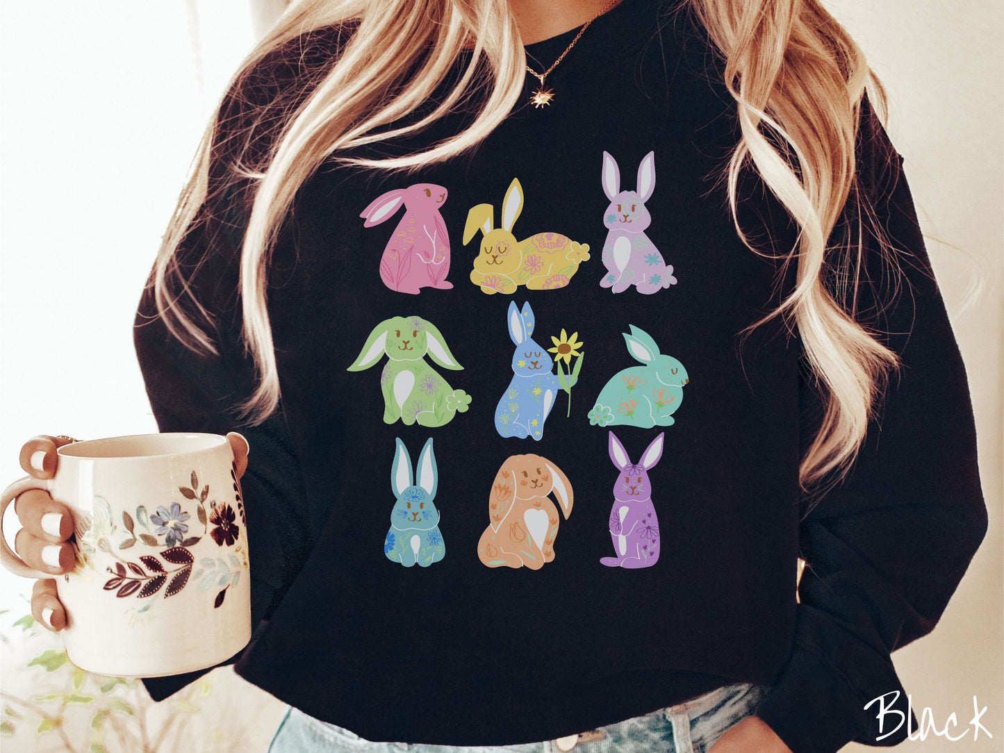 A woman wearing a cute, vintage black colored sweatshirt with a three by three grid of colorful bunny rabbits. They are from left to right pink, yellow, purple, green, blue, seafoam, light blue, orange, and purple with flowers near them.