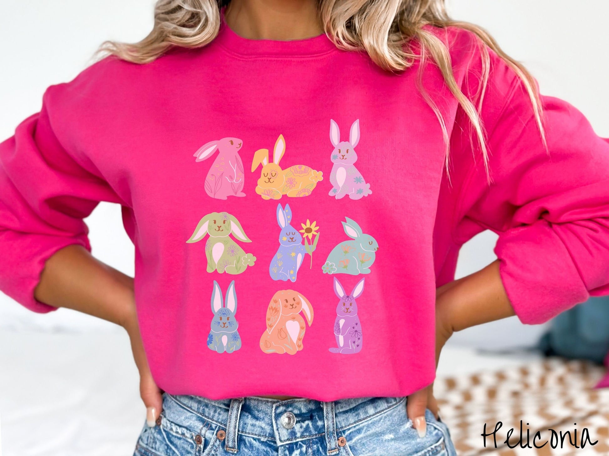 A woman wearing a cute, vintage heliconia colored sweatshirt with a three by three grid of colorful bunny rabbits. They are from left to right pink, yellow, purple, green, blue, seafoam, light blue, orange, and purple with flowers near them.