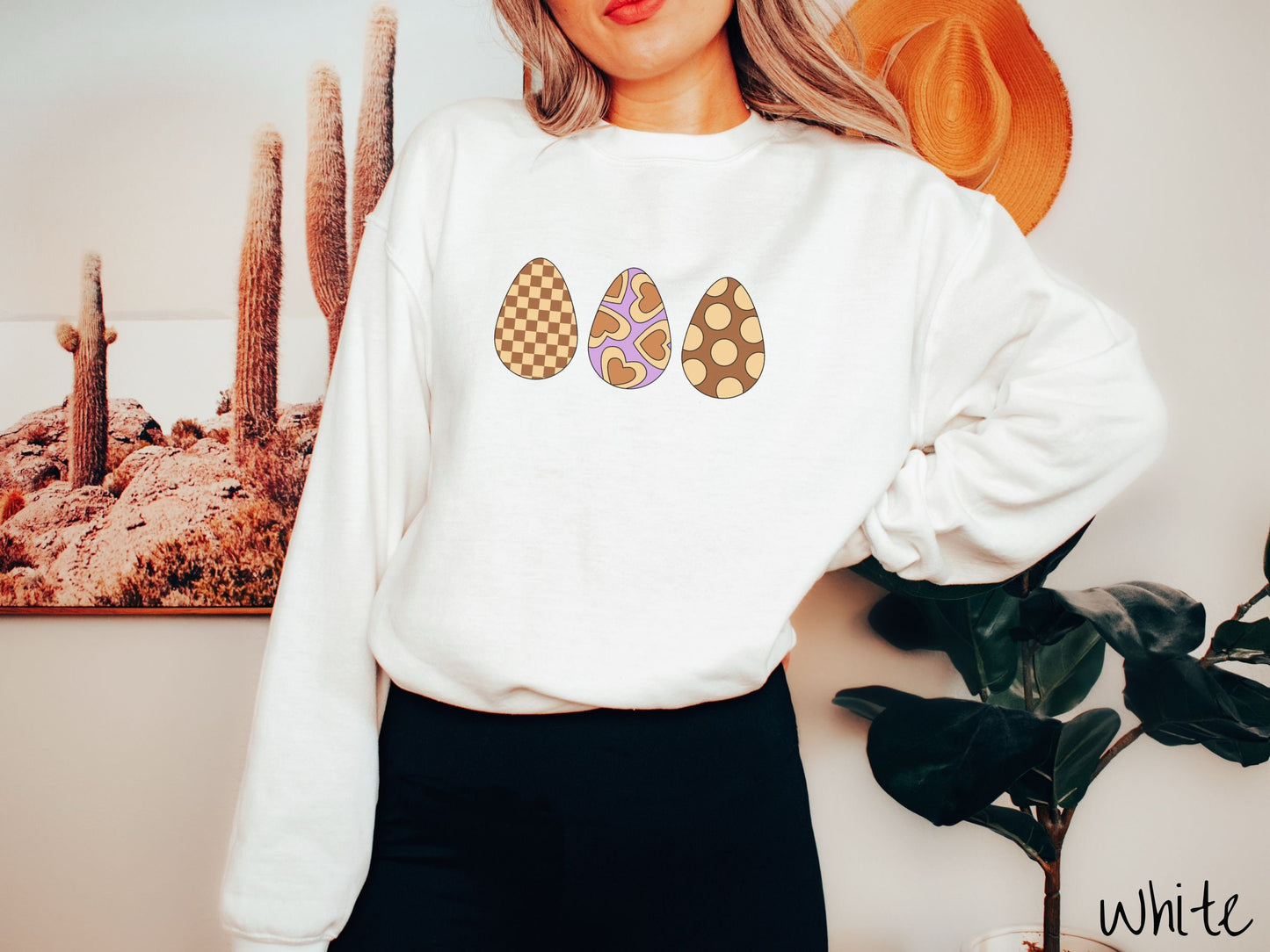 A woman wearing a cute, vintage white colored sweatshirt with three Easter eggs. The left has a light yellow and brown checkerboard pattern, the middle is purple with light yellow brown hearts, and the right is brown with light yellow circles.