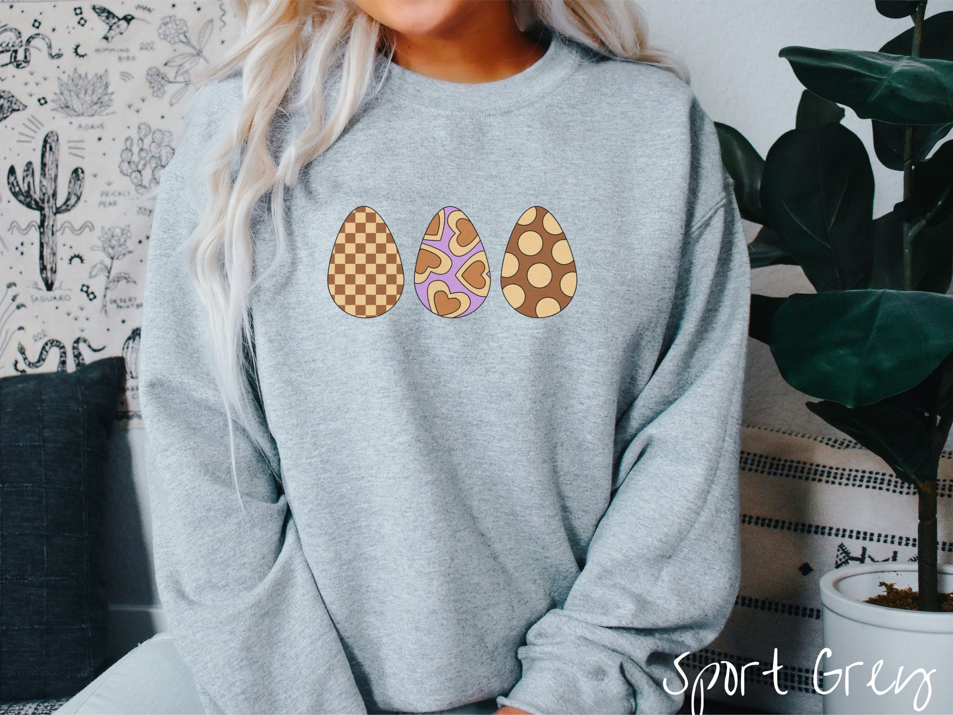 A woman wearing a cute, vintage sport grey colored sweatshirt with three Easter eggs. The left has a light yellow and brown checkerboard pattern, the middle is purple with light yellow brown hearts, and the right is brown with light yellow circles.