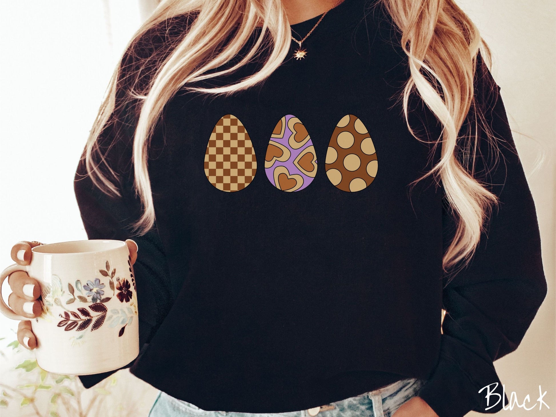 A woman wearing a cute, vintage black colored sweatshirt with three Easter eggs. The left has a light yellow and brown checkerboard pattern, the middle is purple with light yellow brown hearts, and the right is brown with light yellow circles.