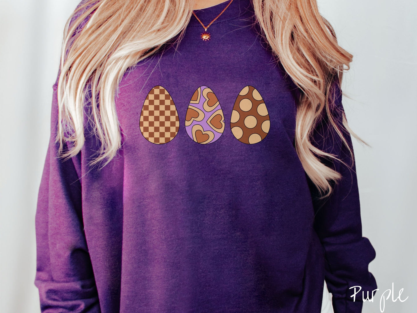 A woman wearing a cute, vintage purple colored sweatshirt with three Easter eggs. The left has a light yellow and brown checkerboard pattern, the middle is purple with light yellow brown hearts, and the right is brown with light yellow circles.