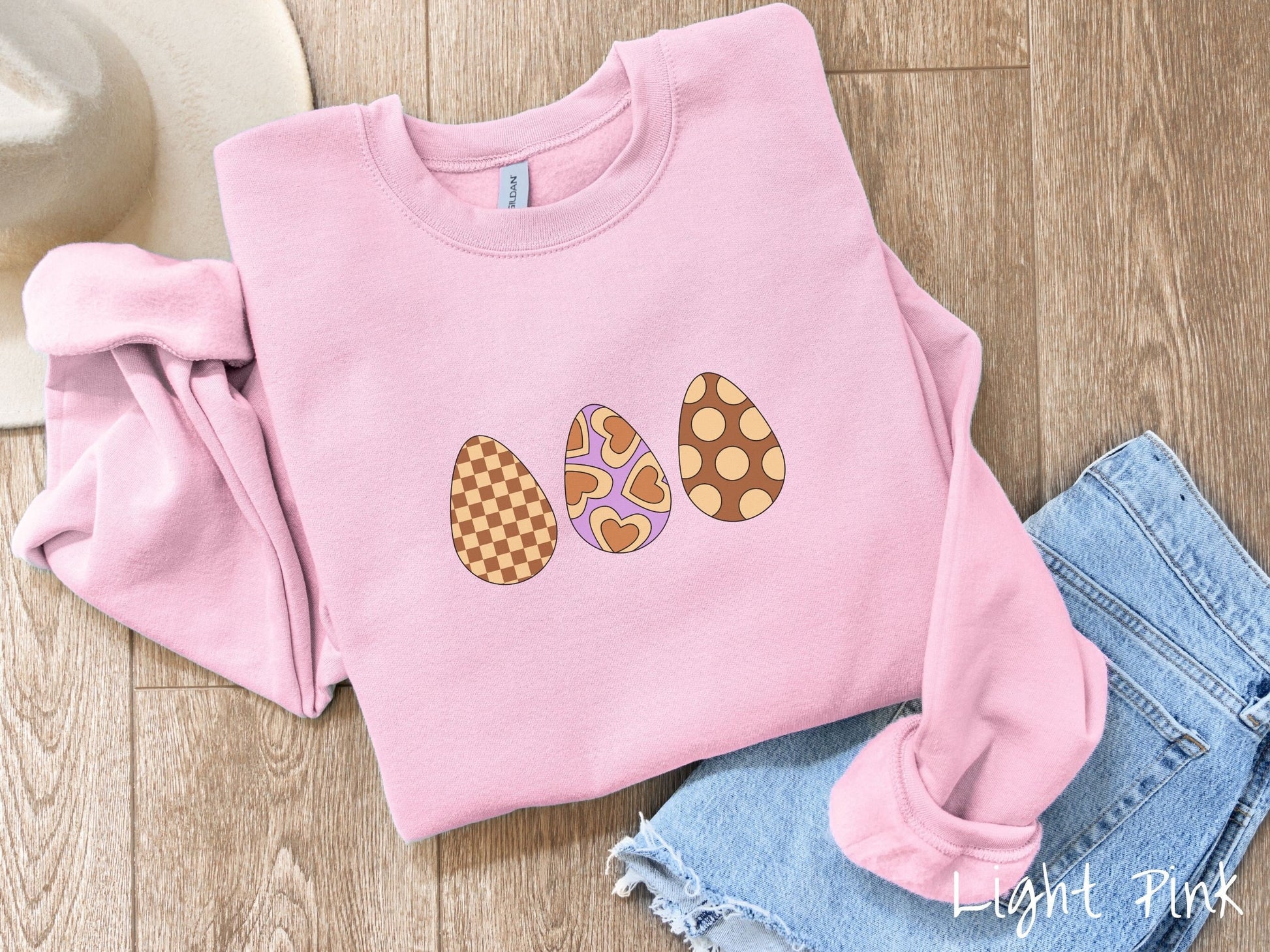A woman wearing a cute, vintage light pink colored sweatshirt with three Easter eggs. The left has a light yellow and brown checkerboard pattern, the middle is purple with light yellow brown hearts, and the right is brown with light yellow circles.