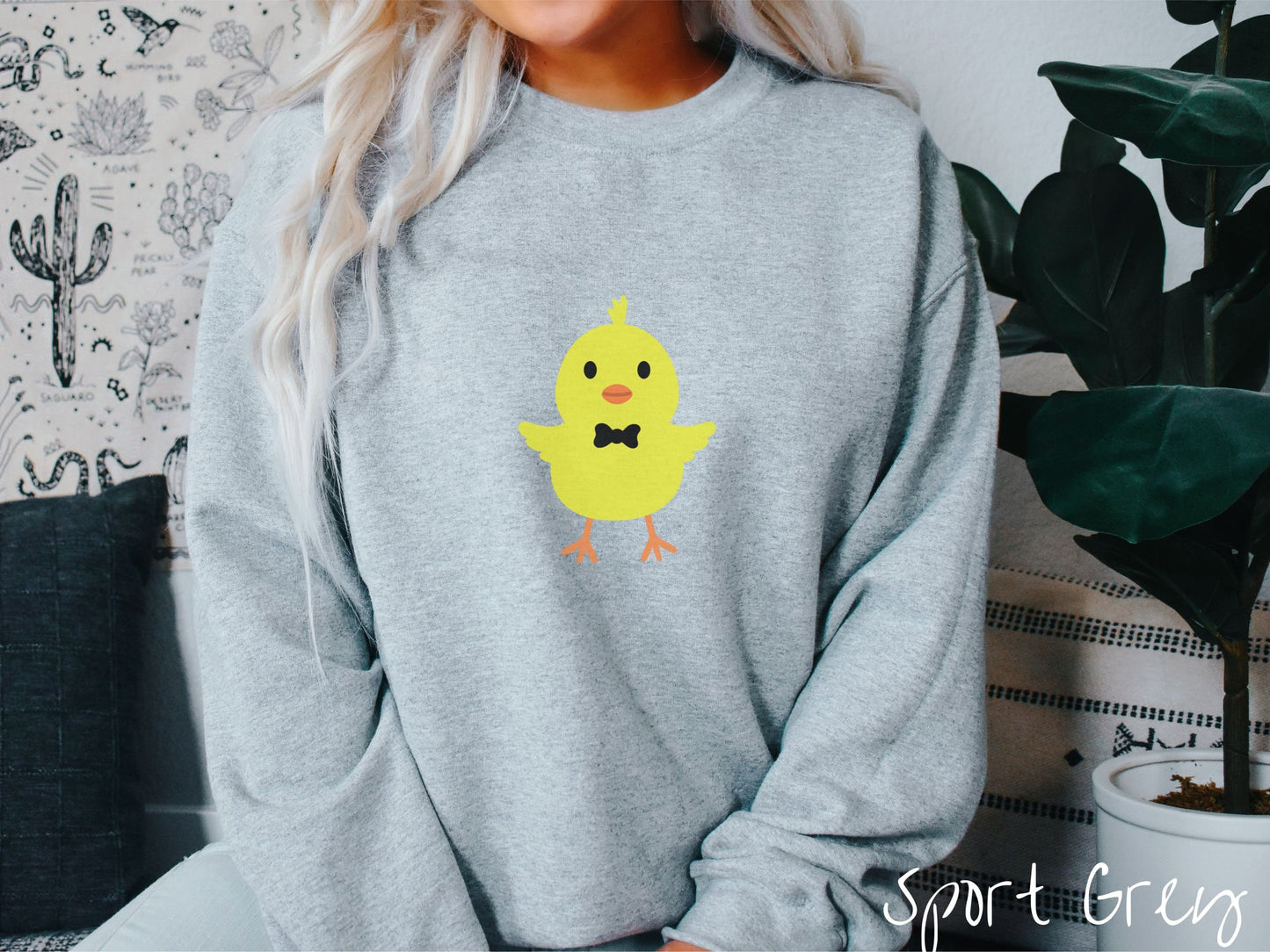 A woman wearing a cute, vintage sport grey colored sweatshirt with a yellow baby chick with a black bowtie and its wings spread is standing and smiling. It has an orange mouth and feet and yellow feathers sticking out the top of its head.