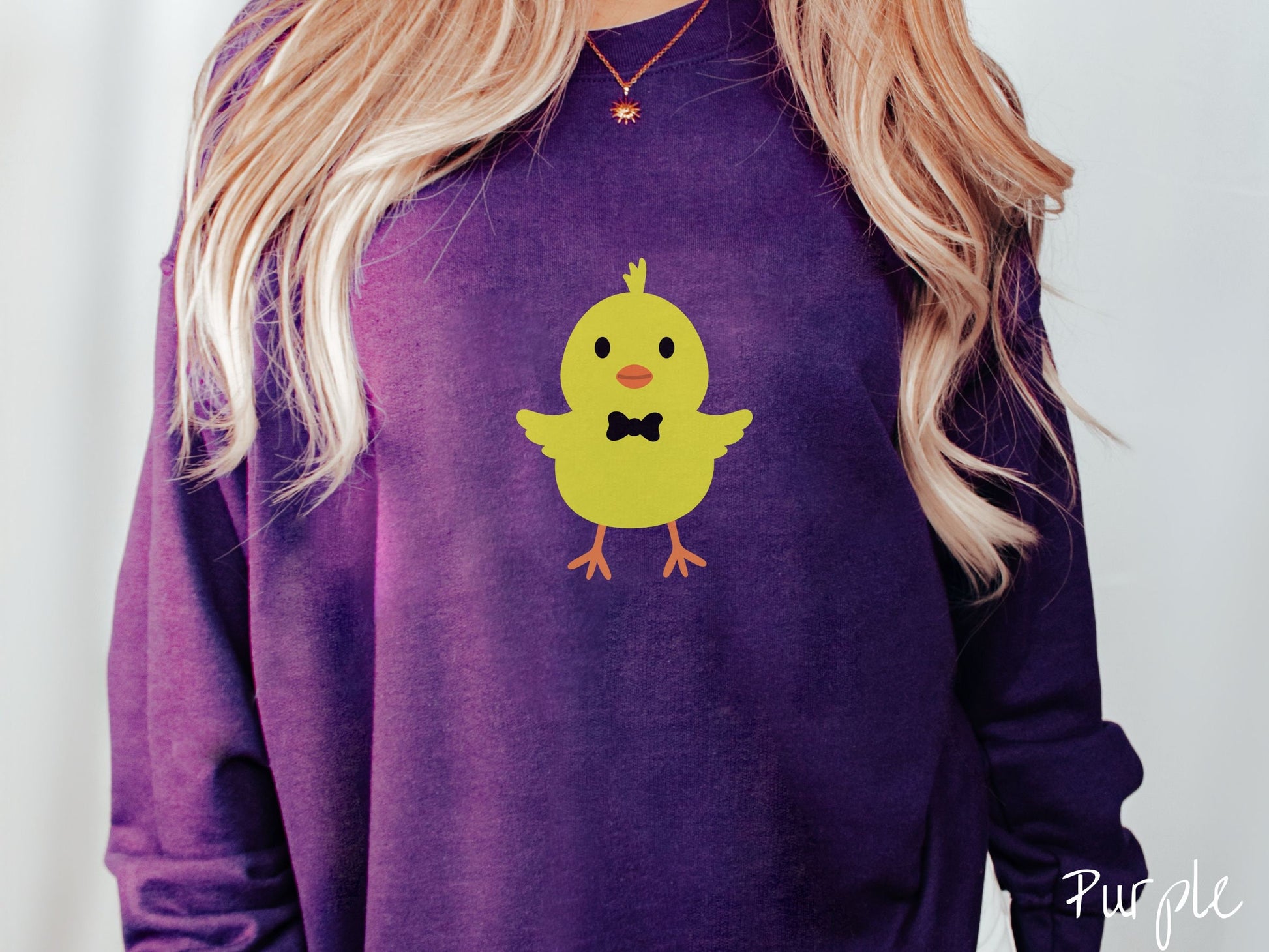 A woman wearing a cute, vintage purple colored sweatshirt with a yellow baby chick with a black bowtie and its wings spread is standing and smiling. It has an orange mouth and feet and yellow feathers sticking out the top of its head.