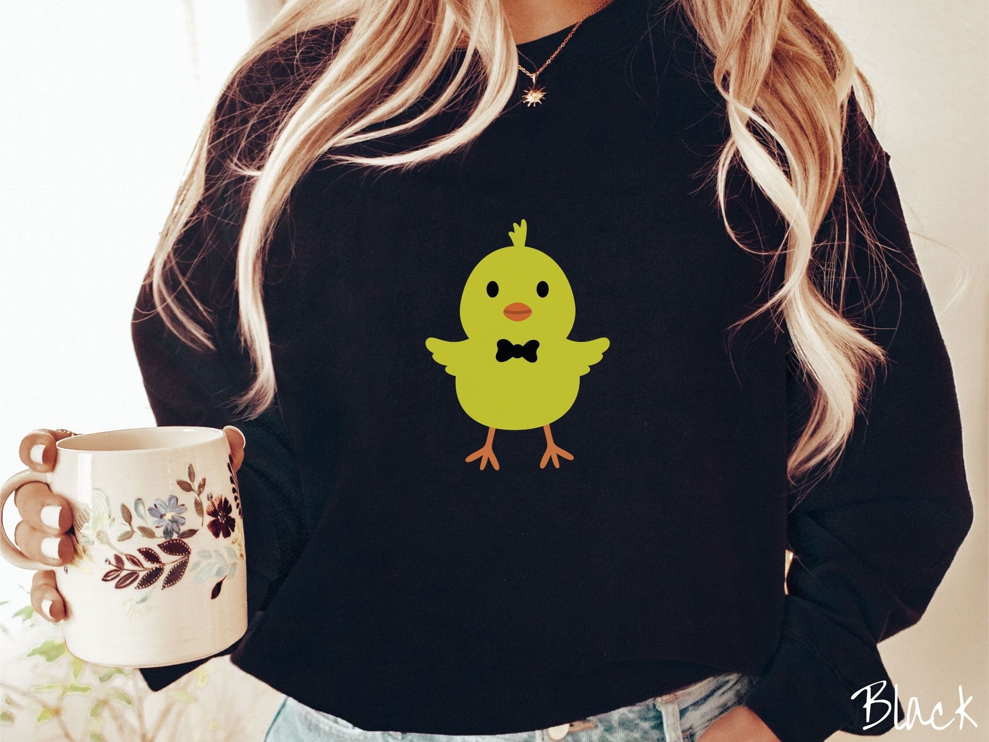 A woman wearing a cute, vintage black colored sweatshirt with a yellow baby chick with a black bowtie and its wings spread is standing and smiling. It has an orange mouth and feet and yellow feathers sticking out the top of its head.