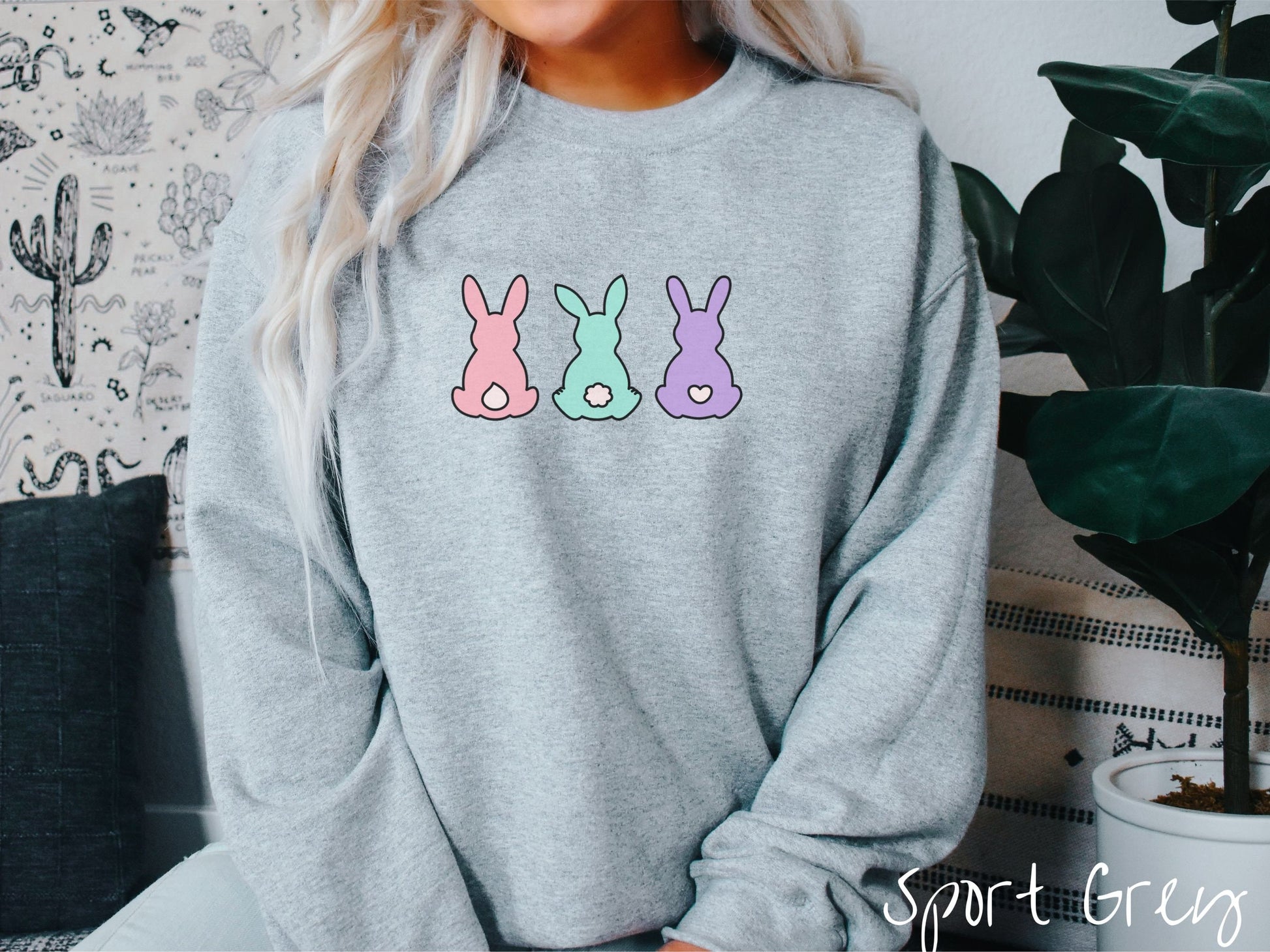 A woman wearing a cute, vintage sport grey colored sweatshirt with three colorful bunny rabbits sitting facing away. One is pink with a white tail, another bay with white tail, and the third is purple with a white, heart-shaped tail.