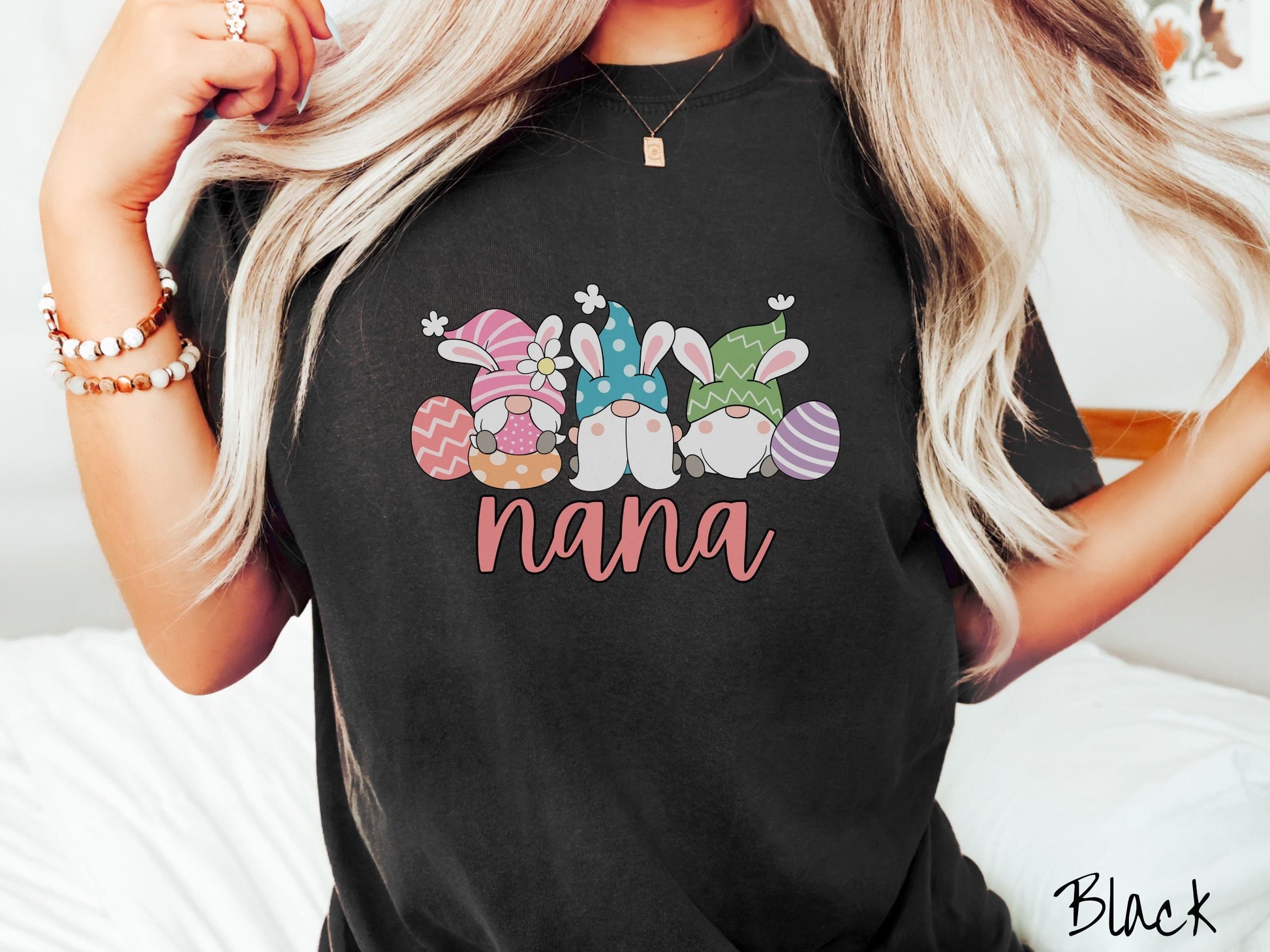 A woman wearing a cute, vintage black colored shirt with text Nana below three gnomes with rabbit ears sitting next to each other. The left one is wearing pink, the middle blue, and right in green, with colorful easter eggs scattered about.