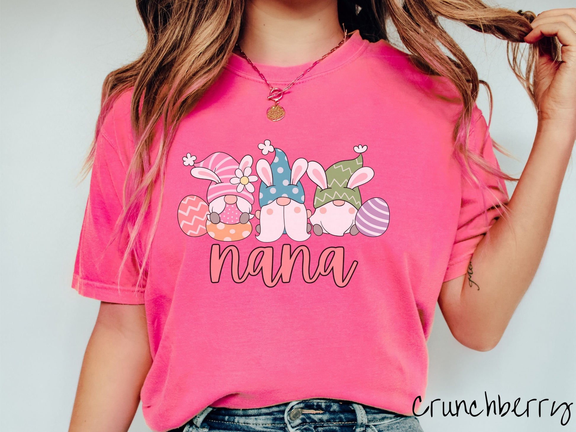 A woman wearing a cute, vintage crunchberry colored shirt with text Nana below three gnomes with rabbit ears sitting next to each other. The left one is wearing pink, the middle blue, and right in green, with colorful easter eggs scattered about.