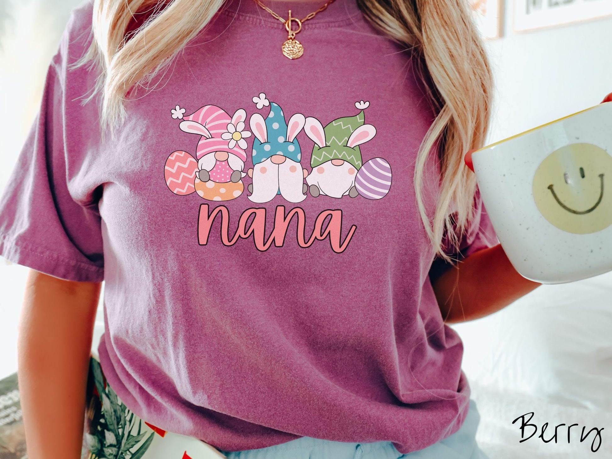 A woman wearing a cute, vintage berry colored shirt with text Nana below three gnomes with rabbit ears sitting next to each other. The left one is wearing pink, the middle blue, and right in green, with colorful easter eggs scattered about.