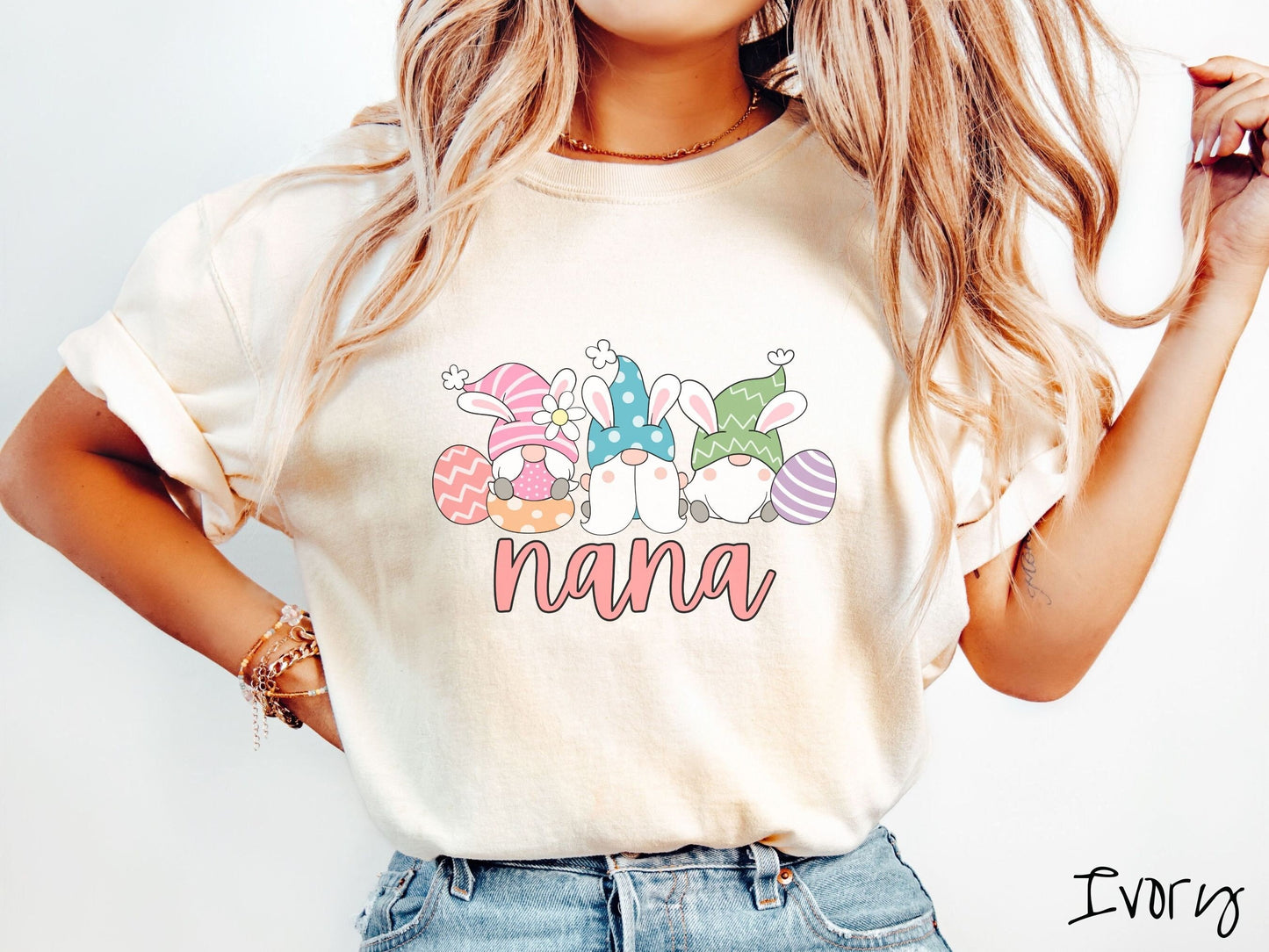 A woman wearing a cute, vintage ivory colored shirt with text Nana below three gnomes with rabbit ears sitting next to each other. The left one is wearing pink, the middle blue, and right in green, with colorful easter eggs scattered about.