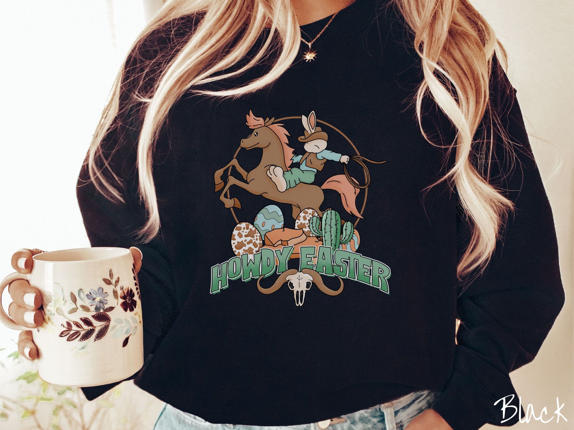 A woman wearing a cute, vintage black colored sweatshirt with a cowboy rabbit riding a bucking, brown horse while holding a lasso rope. Below is green text Howdy Easter along with colorful Easter eggs, a green cactus, and a horned bull skull.