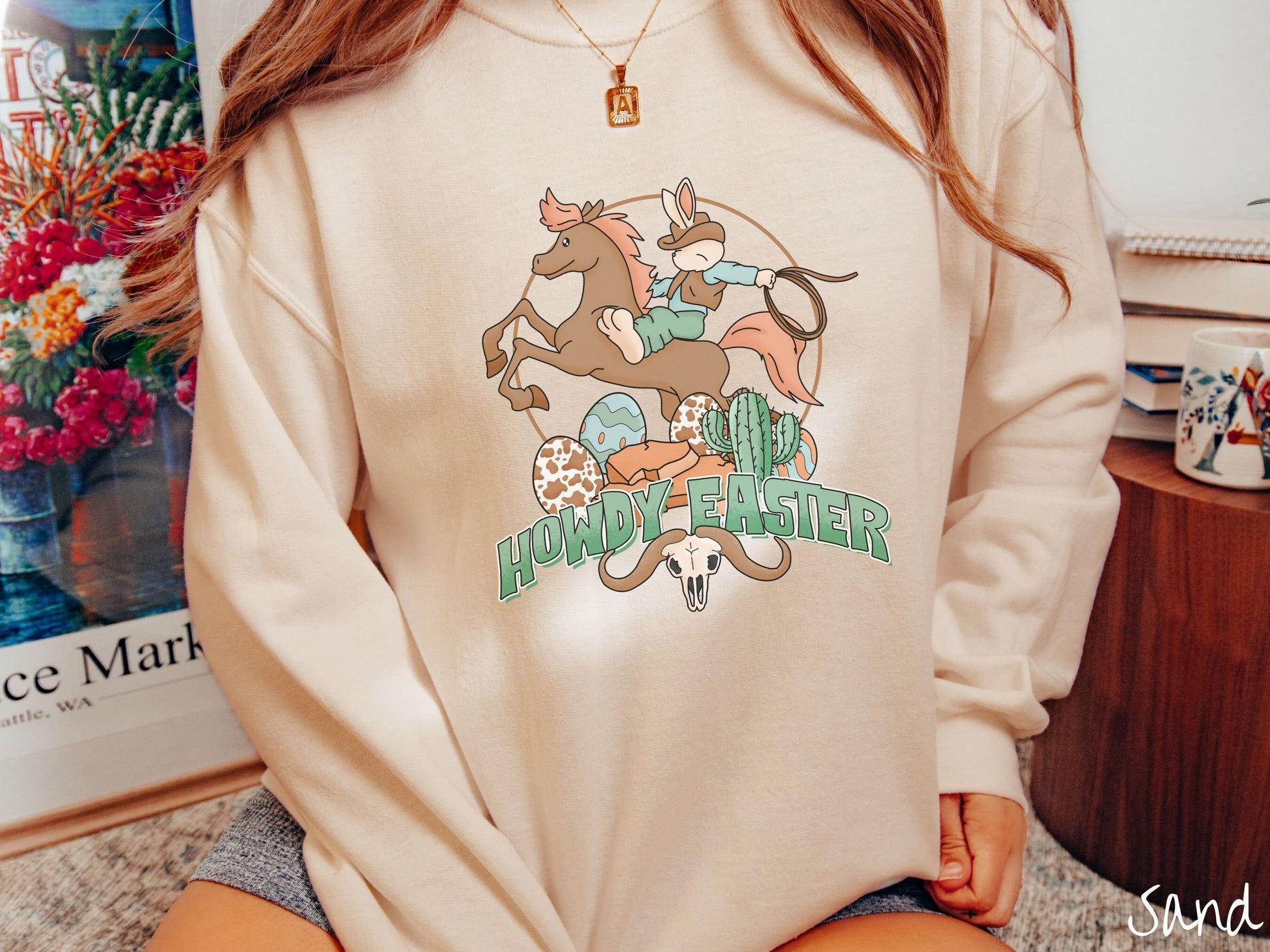 A woman wearing a cute, vintage sand colored sweatshirt with a cowboy rabbit riding a bucking, brown horse while holding a lasso rope. Below this is the green text Howdy Easter along with colorful Easter eggs, a green cactus, and a horned bull skull.