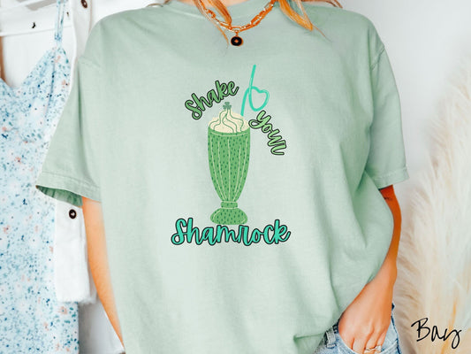 A woman wearing a vintage, bay colored shirt with the multicolor text Shake Your Shamrock and between this saying is a green glass filled with a vanilla milkshake with green syrup and a green twisty straw.