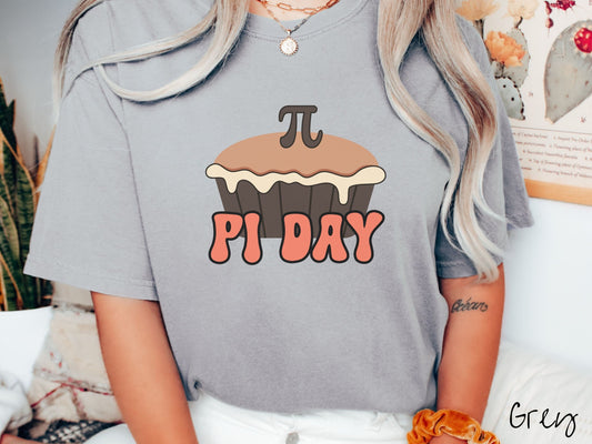 A woman wearing a cute, vintage grey colored comfort colors shirt with the text Pi Day in orange font, above that is a brown pumpkin pie with a pi symbol on the top of the pie.