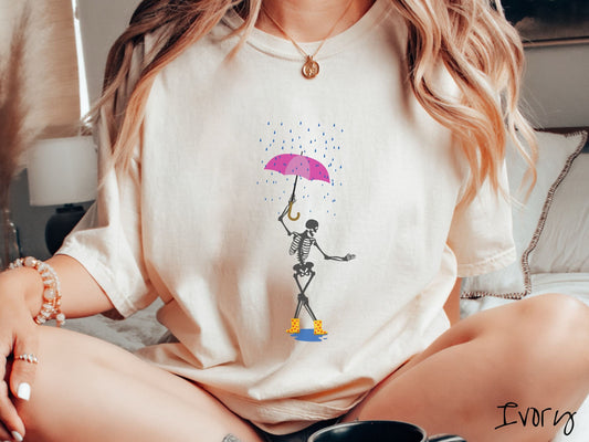 A woman wearing a cute ivory colored shirt with a dark grey female skeleton dancing in the rain with a pink and purple umbrella and wearing yellow with red polka dot rain boots.