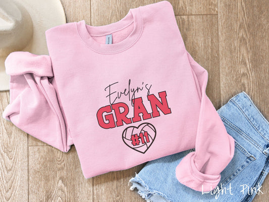 A woman wearing a cute light pink colored sweatshirt with black and red text saying Evelyns Gran. Below is a black, heart-shaped volleyball outline with #11 in red font written across it.