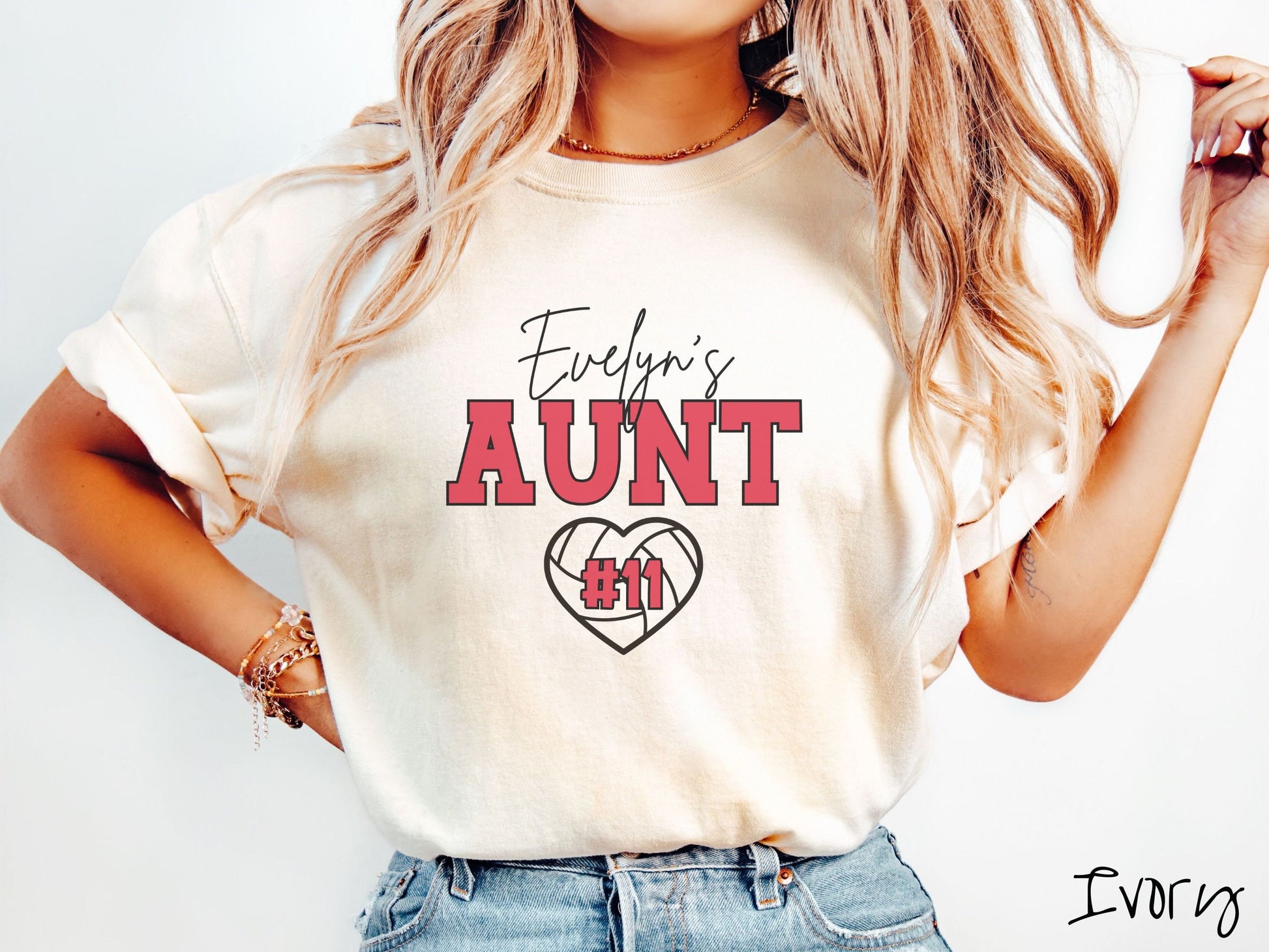 A woman wearing a cute ivory colored t-shirt with black and red text saying Evelyns Aunt. Below is a black, heart-shaped volleyball outline with #11 in red font written across it.