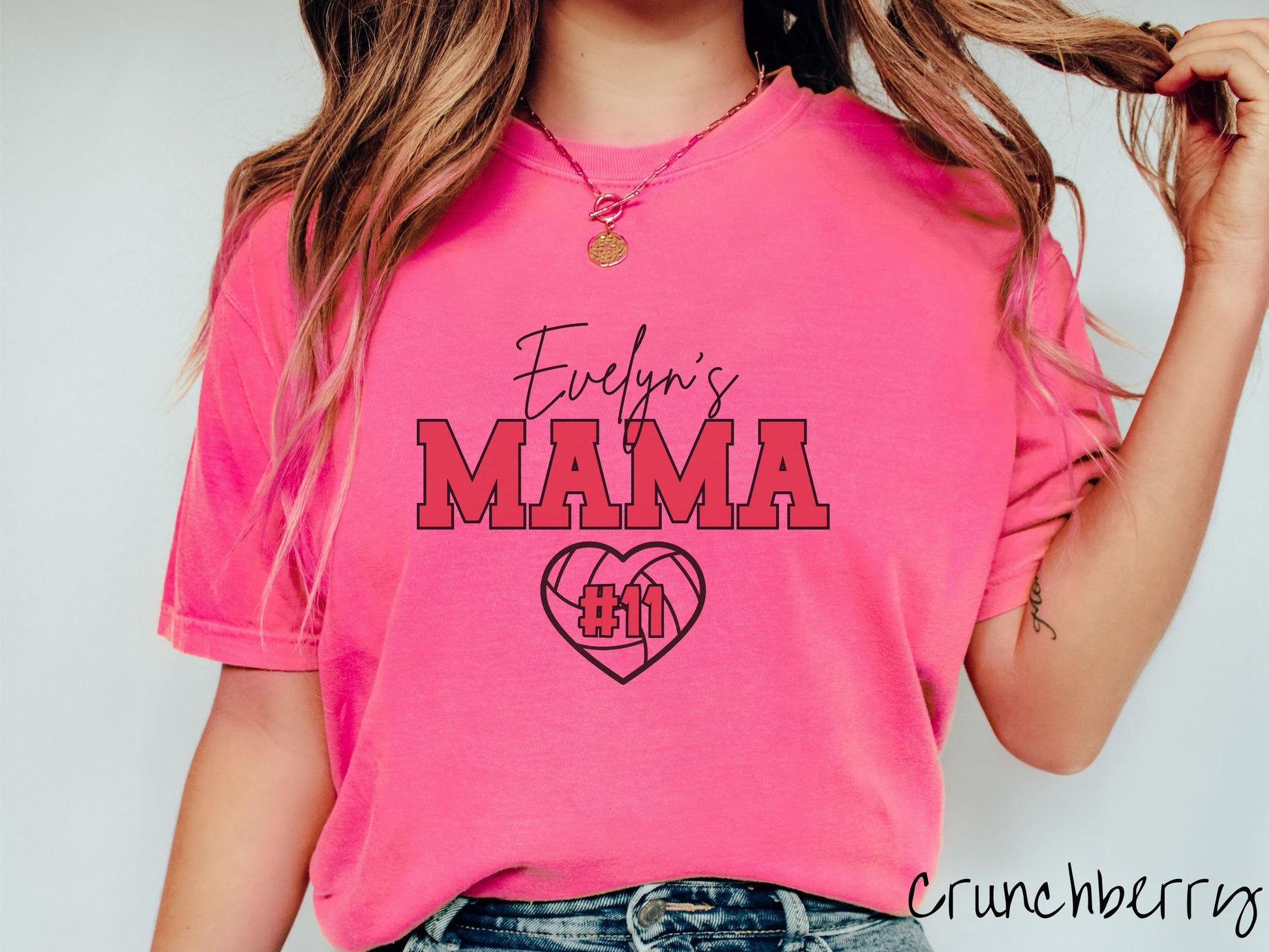 A woman wearing a cute crunchberry colored t-shirt with black and red text saying Evelyns Mama. Below is a black, heart-shaped volleyball outline with #11 in red font written across it.