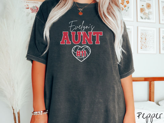 A woman wearing a cute pepper colored t-shirt with white and red text saying Evelyns Aunt. Below is a white, heart-shaped volleyball outline with #11 in red font written across it.