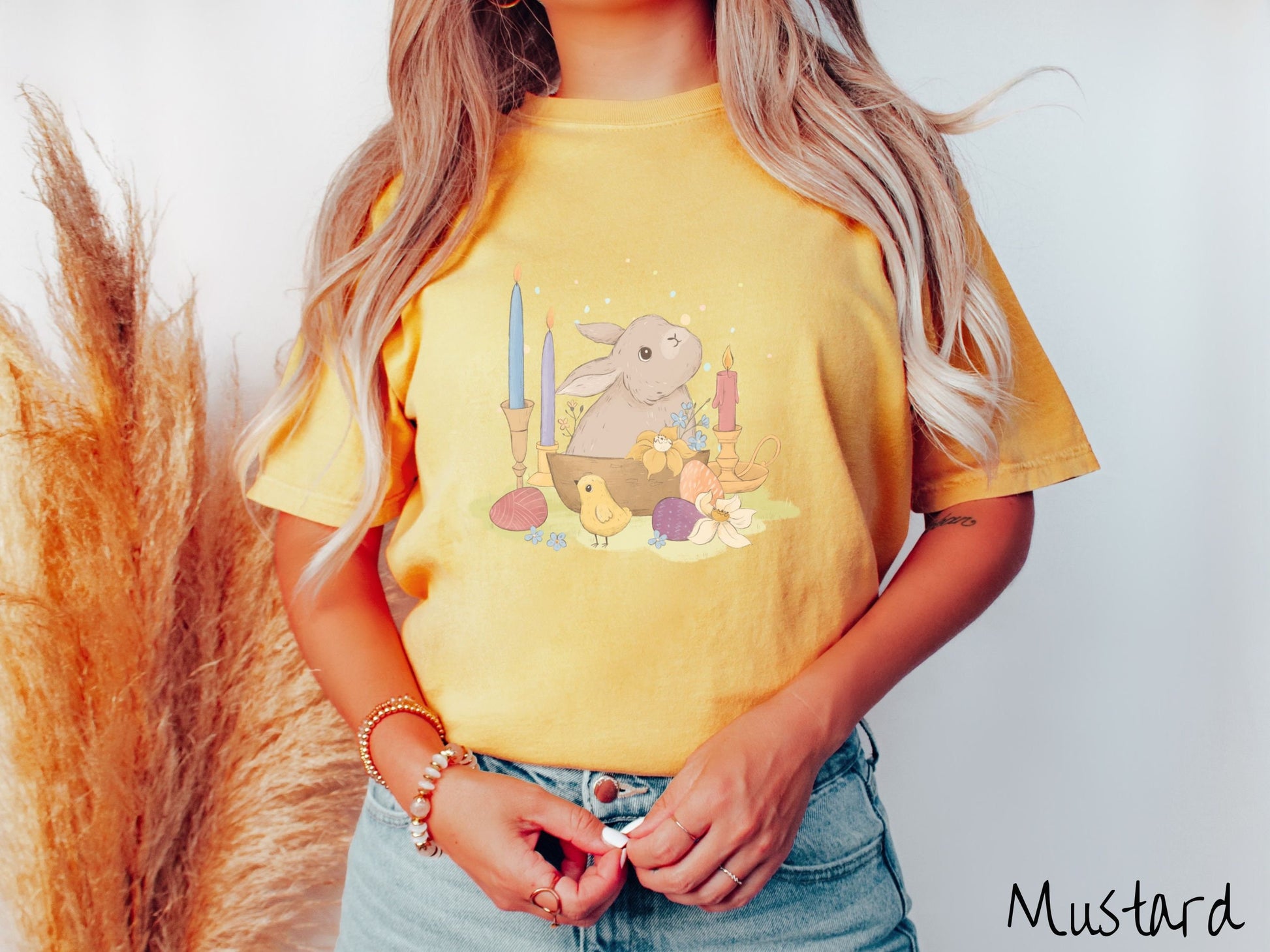 A woman wearing a cute, vintage mustard colored shirt with a gray bunny rabbit sitting in between three lit candles, some red, orange, and purple eggs, and yellow and white flowers.