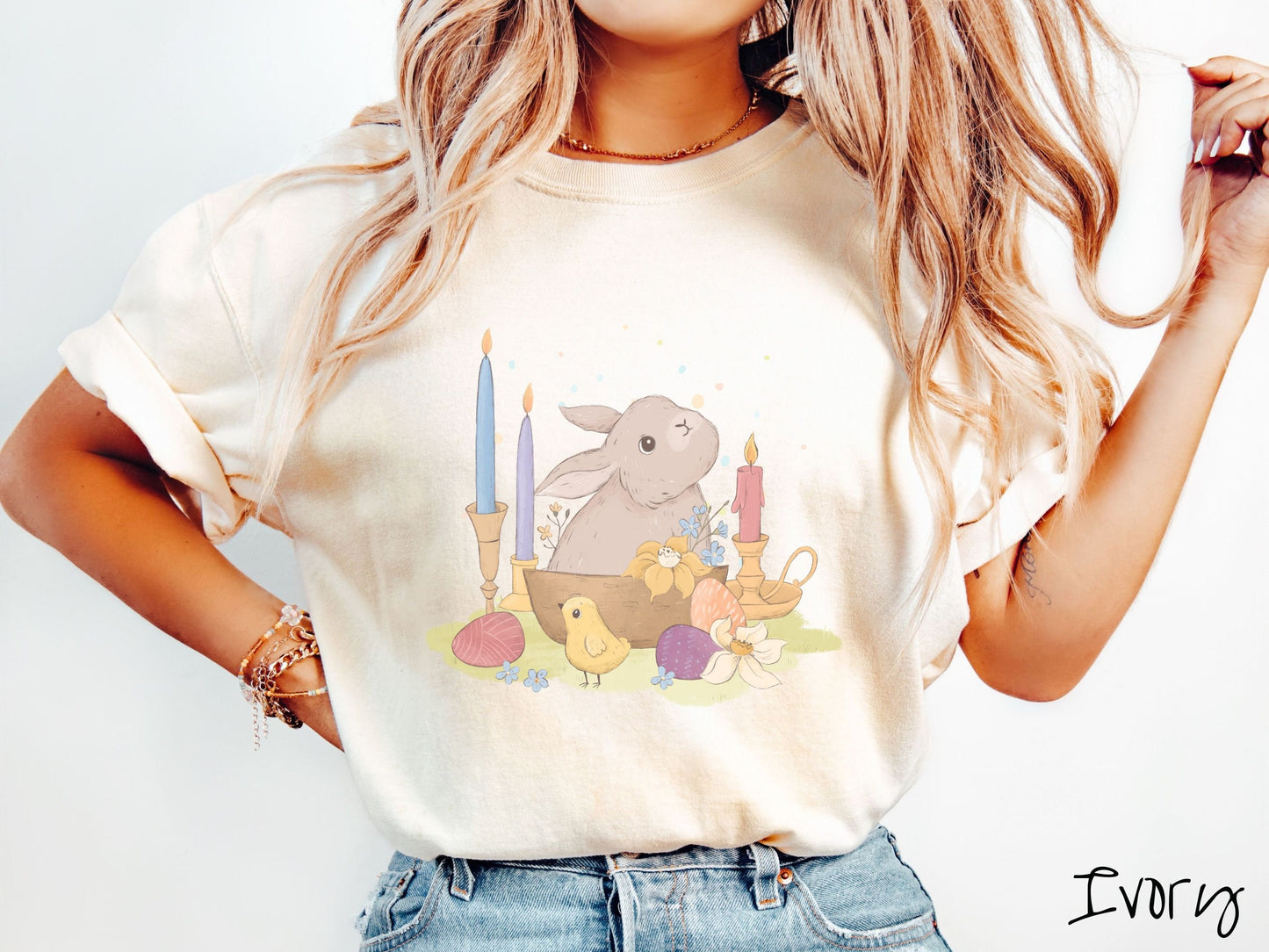 A woman wearing a cute, vintage ivory colored shirt with a gray bunny rabbit sitting in between three lit candles, some red, orange, and purple eggs, and yellow and white flowers.