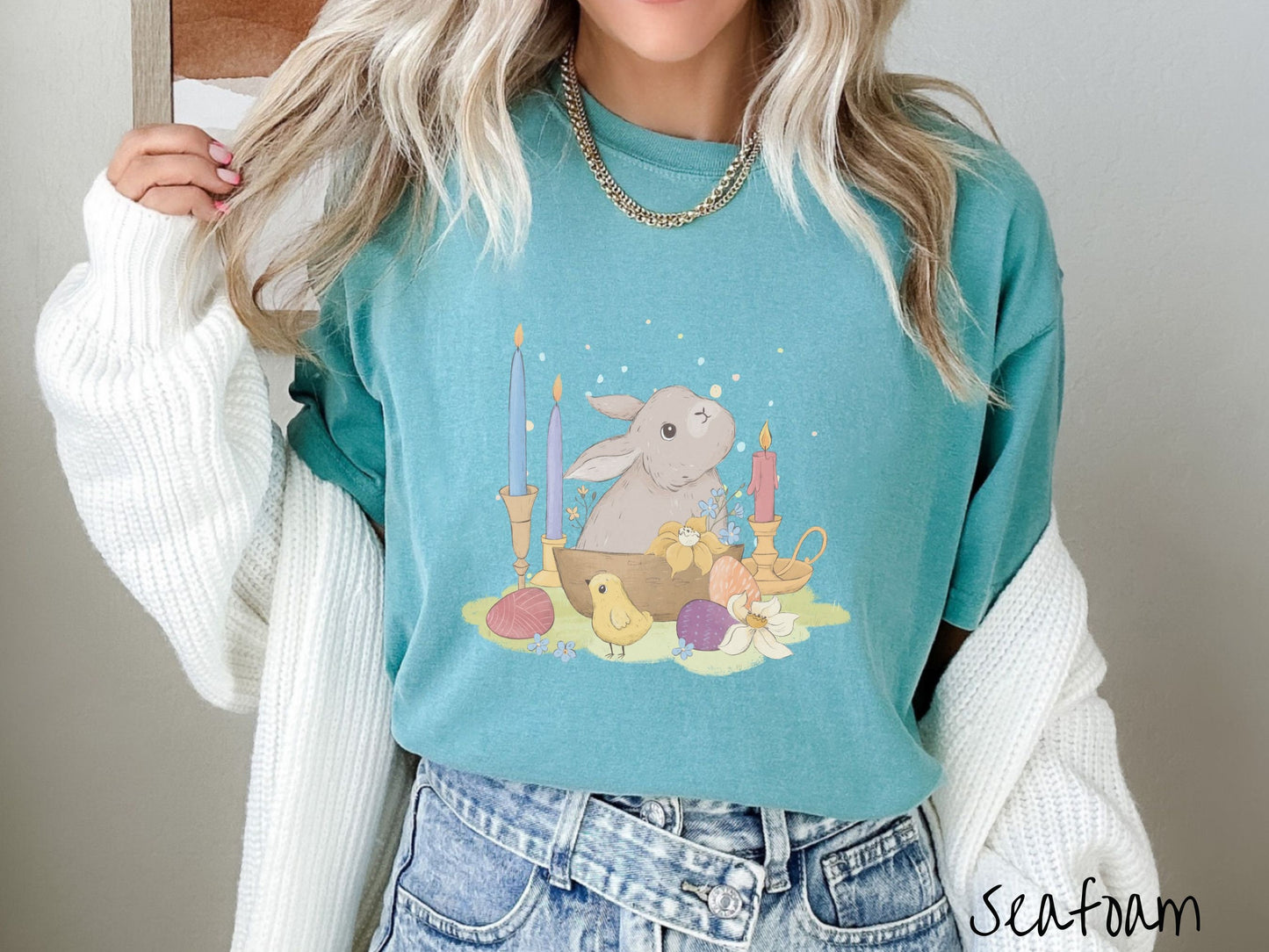 A woman wearing a cute, vintage seafoam colored shirt with a gray bunny rabbit sitting in between three lit candles, some red, orange, and purple eggs, and yellow and white flowers.