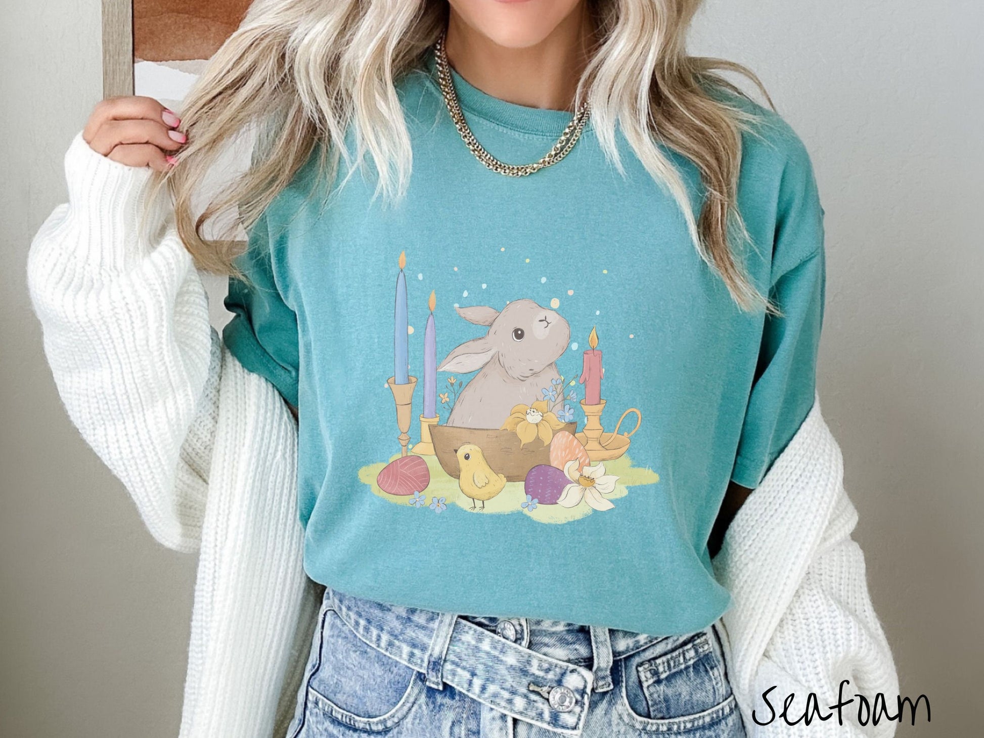 A woman wearing a cute, vintage seafoam colored shirt with a gray bunny rabbit sitting in between three lit candles, some red, orange, and purple eggs, and yellow and white flowers.