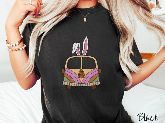 A woman wearing a cute, vintage black colored shirt with an old-fashioned, hippie style van with pastel-colored stripes on the front and light orange paint covering the rest. The van has a brown egg on the front and two, floppy bunny ears on top.