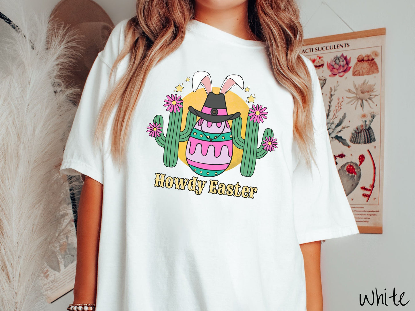 A woman wearing a cute, vintage white colored shirt with an Easter egg with green, pink and purple stripes, wearing a black cowboy hat and white bunny ears, and it’s standing in between two green cacti with pink flowers in front of the sun.