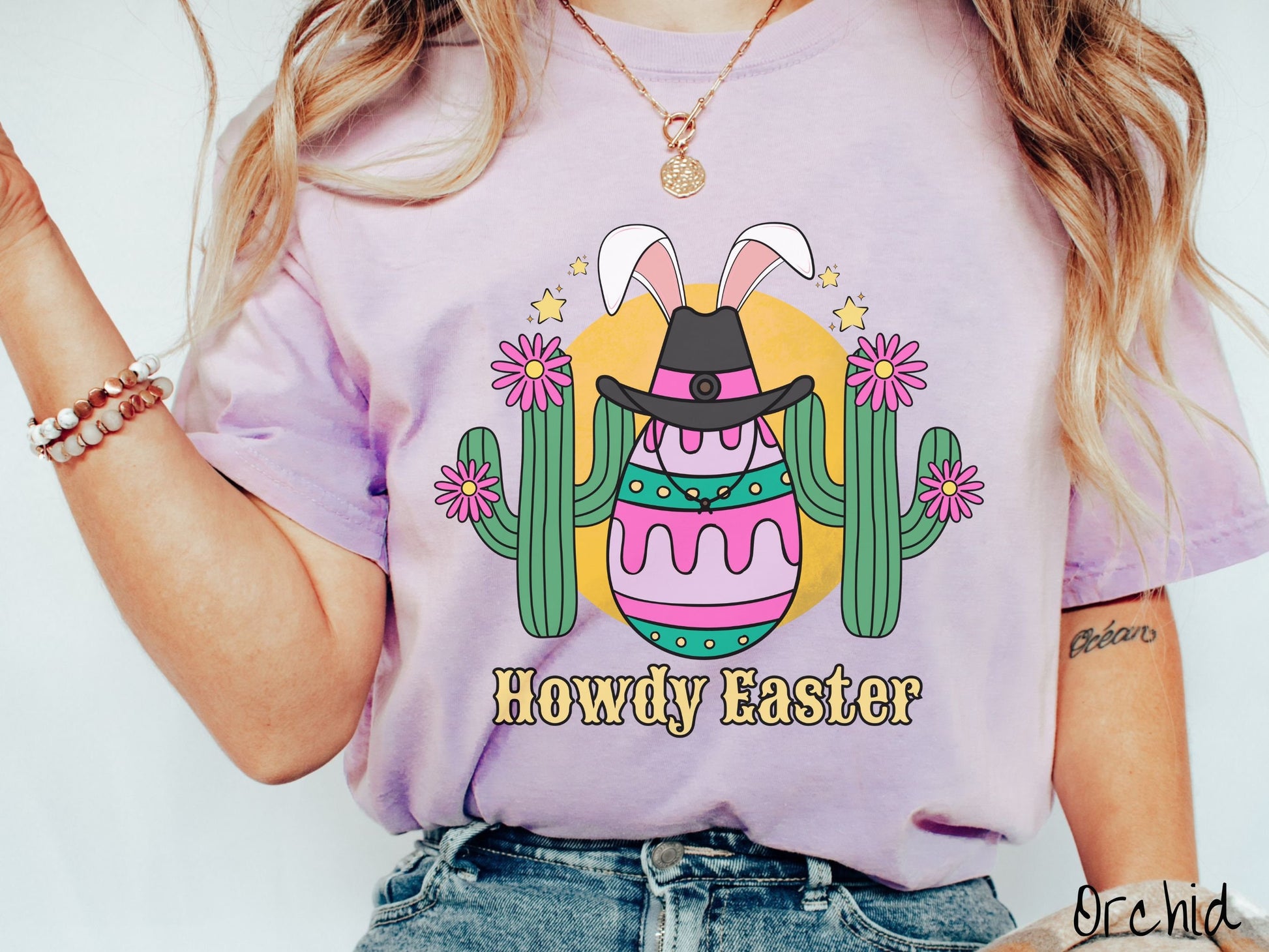 A woman wearing a cute, vintage orchid colored shirt with an Easter egg with green, pink and purple stripes, wearing a black cowboy hat and white bunny ears, and it’s standing in between two green cacti with pink flowers in front of the sun.