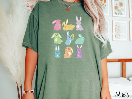 A woman wearing a cute, vintage moss colored shirt with a three by three grid of colorful bunny rabbits. They are from left to right pink, yellow, purple, green, blue, seafoam, light blue, orange, and purple and have flowers and plants drawn on them.