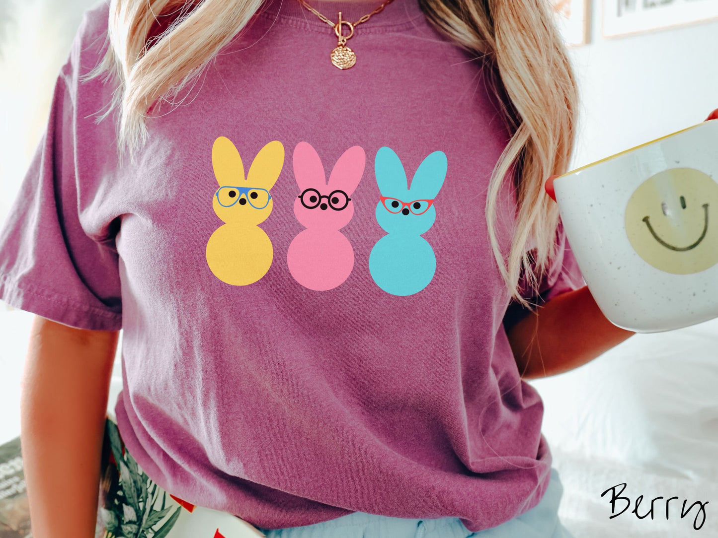 A woman wearing a cute, vintage berry colored shirt with three peep-like rabbits on the front. One is yellow and wearing blue glasses, the middle is pink with black glasses, and the one on the right is light blue with red glasses.