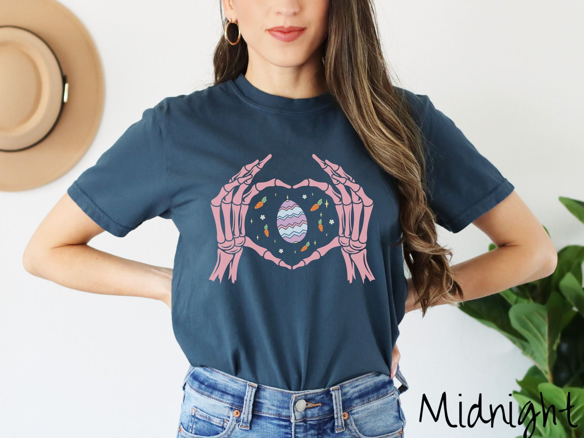 A woman wearing a cute, vintage midnight colored shirt with two skeleton hands forming a heart shape, and within the heart are an orange, purple, and blue striped Easter egg, carrots with green leaves, tiny flowers, and twinkling stars.