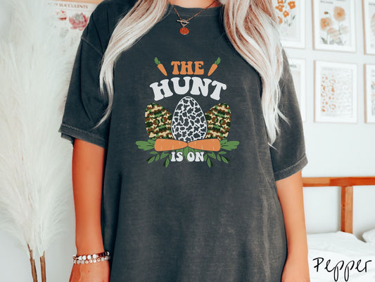 A woman wearing a cute, vintage pepper colored shirt with the text The Hunt Is On in orange and white font, with three Easter eggs painted in woodland camo and black and white camo in front. Above and below the eggs are carrots with leafy ends.