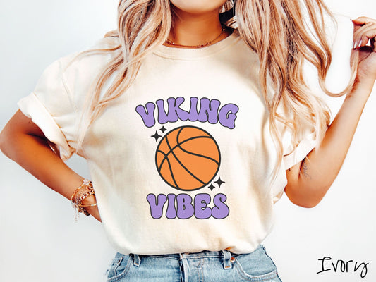 A woman wearing a cute ivory colored shirt with purple text saying Viking Vibes. There is an orange basketball with black twinkling stars around it between the text.