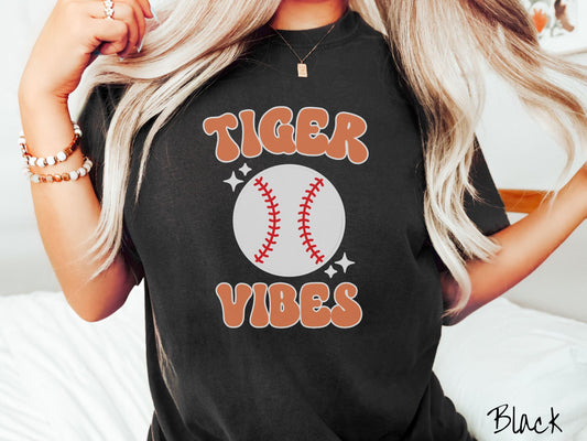 A woman wearing a cute black colored shirt with orange text saying Tiger Vibes. There is a white baseball with red stitching between the text with white twinkling stars.