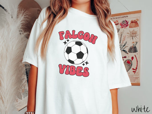 A woman wearing a cute white colored shirt with red text saying Falcon Vibes. There is a black and white checkered soccer ball with black twinkling stars around it between the text.